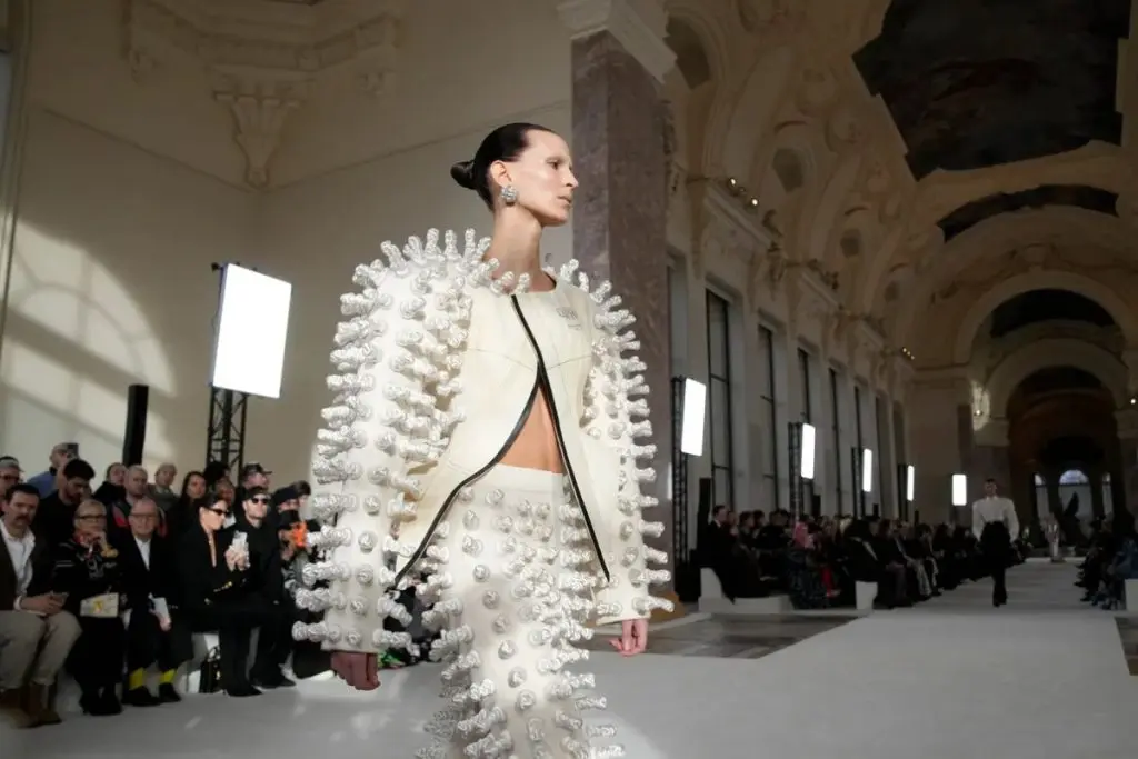 Model walking the runway at a Paris fashion show wearing a sculptural white textured outfit in a historic venue