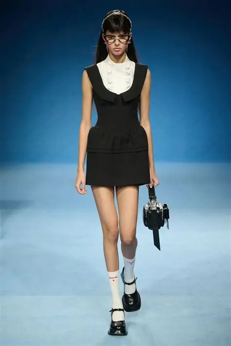 Model walking the runway in a black mini dress with white bib collar and camera bag.